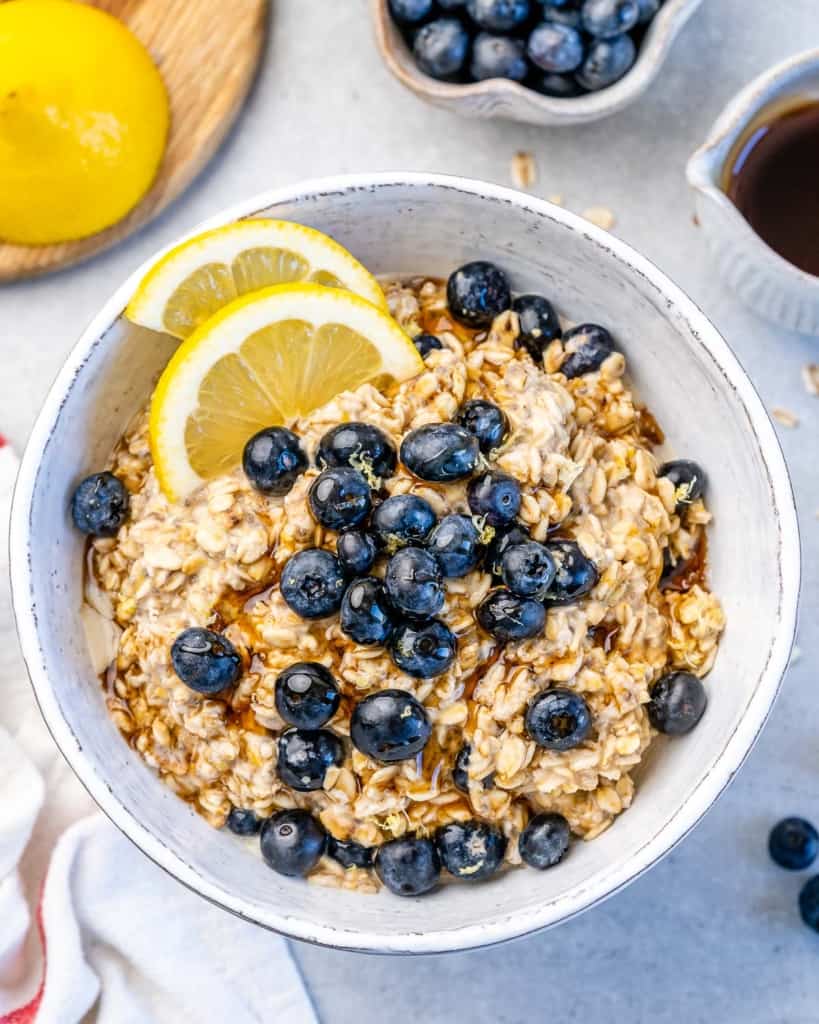 top view of overnight oatmeal in a white bowl topped with blueberries and maple syrup with 2 lemon slices to garnish