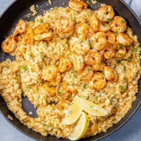 top view risotto in black skillet topped with shrimp in a black skillet with 3 lemon wedges to garnish