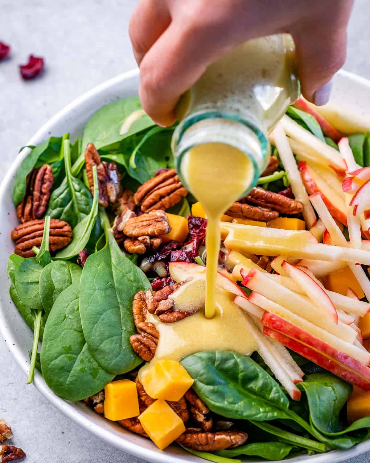 drizzling maple vinaigrette over bowl of spinach salad with apples and pecans