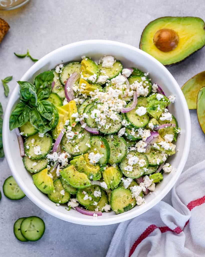 top view of cucumber feta salad in a white bowl and a side of half fresh avocado