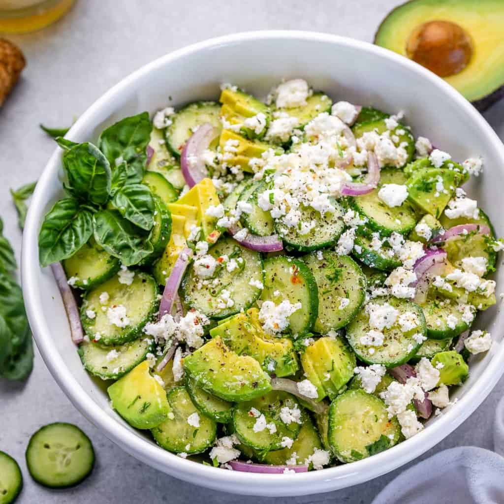 side shot of cucumber salad in a white bowl, topped with feta cheese and fresh basil garnish on the left side of the bowl