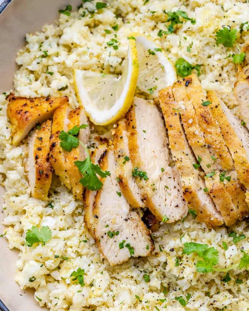 close up sliced chicken breast over cauliflower rice in an orange skillet with cilantro garnishes and lemon slices 