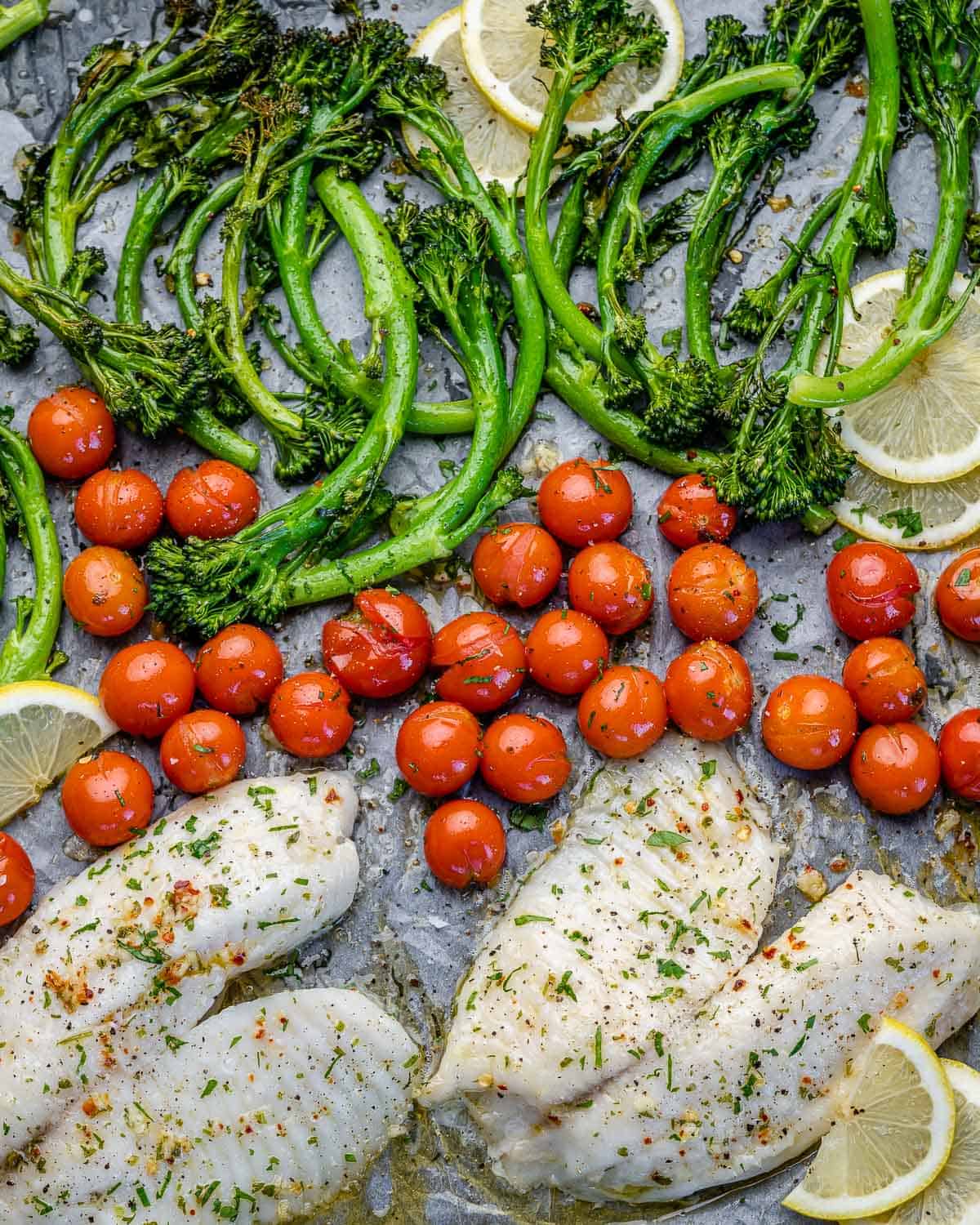 top view of tilapia fillets on the sheet pan with lemons, tomatoes, and broccolini 