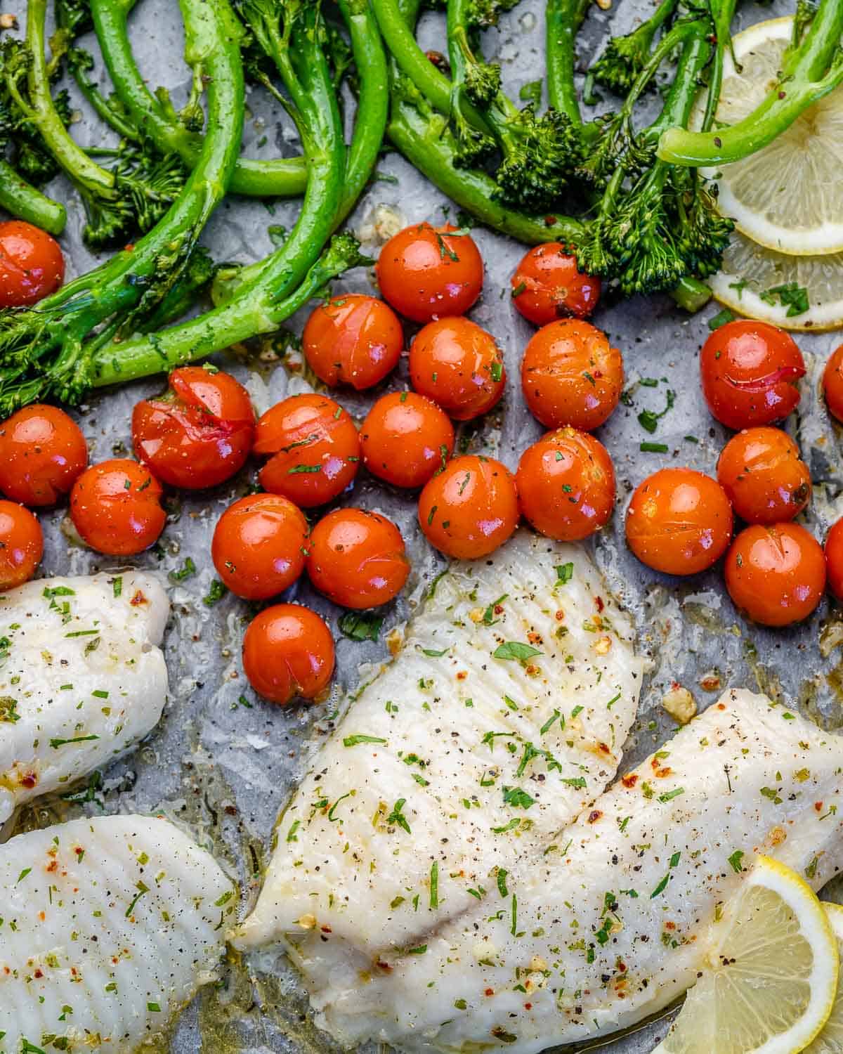 top view of baked tilapia on sheet pan with tomatoes and broccolini with lemon wedges