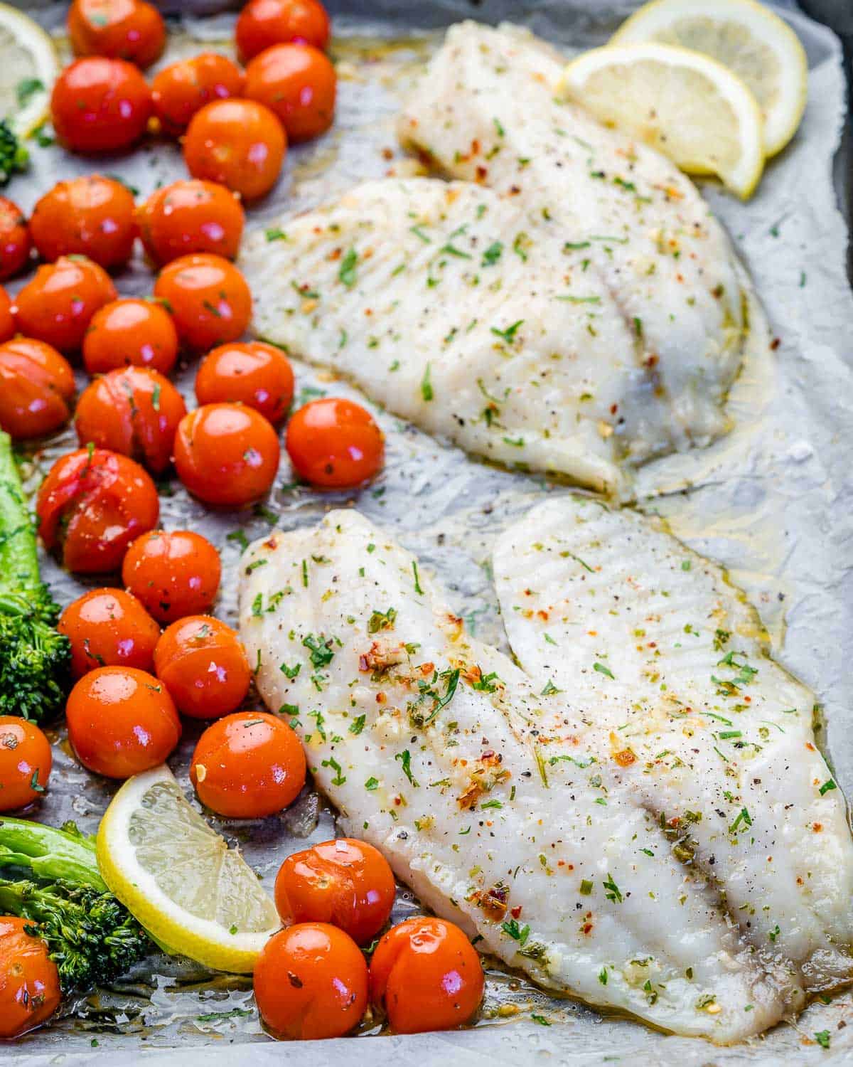 four tilapia fish fillets on sheet pan with tomatoes and lemon