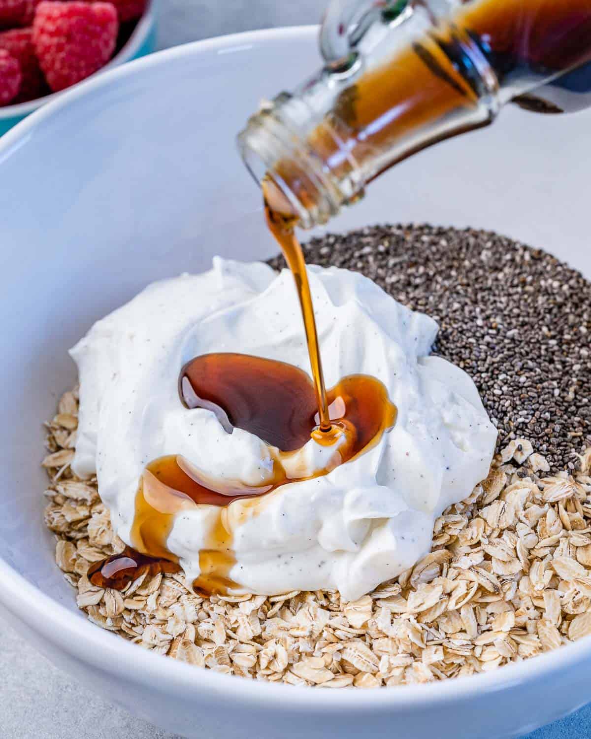 maple syrup being poured over yogurt, oats, and chia seeds in a white bowl.