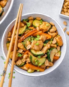 top view chicken and veggie stir fry in a white bowl with two brown chop sticks sitting on the bowl