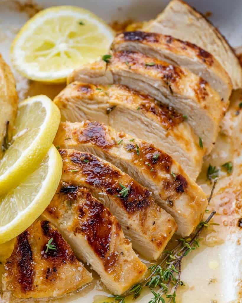 close up sliced roasted chicken breast with sliced lemon garnishes in a white dish