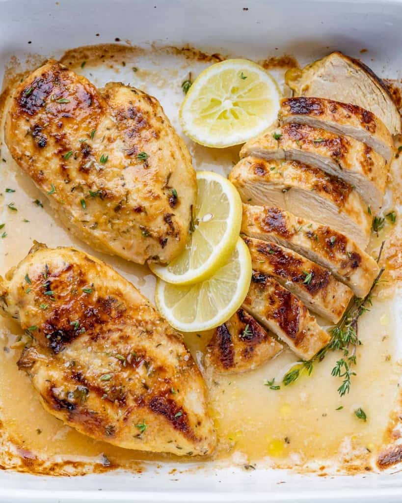 top view roasted chicken breast in a white dish with lemon garnished. Top right chicken breast is sliced
