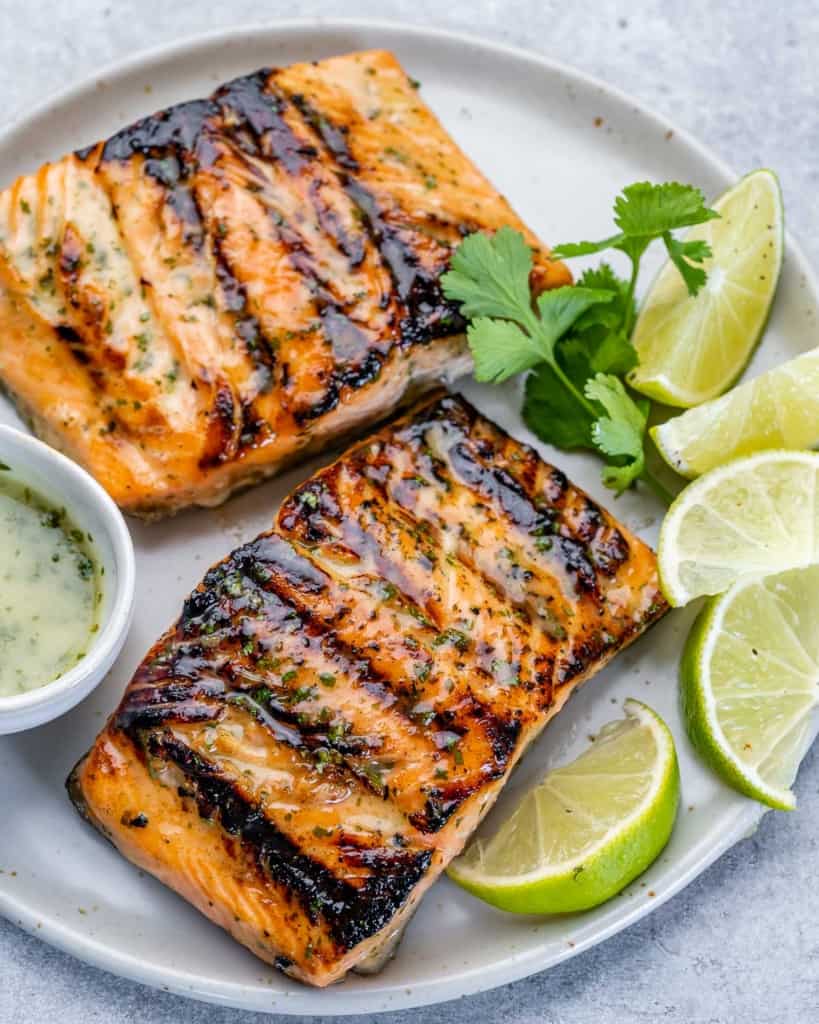 tope view of 2 grilled salmon fillets on a white plate with lime slices to garnish