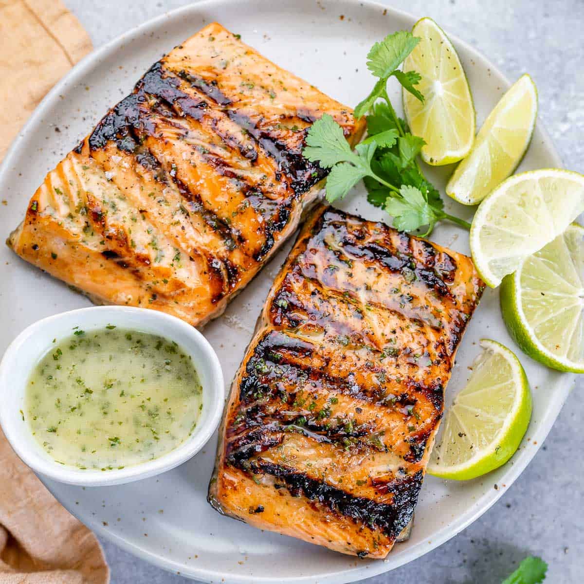 top close up view of 2 grilled salmon filets on a white plate with a side of lime garnished and cilantro lime sauce in a small white bowl