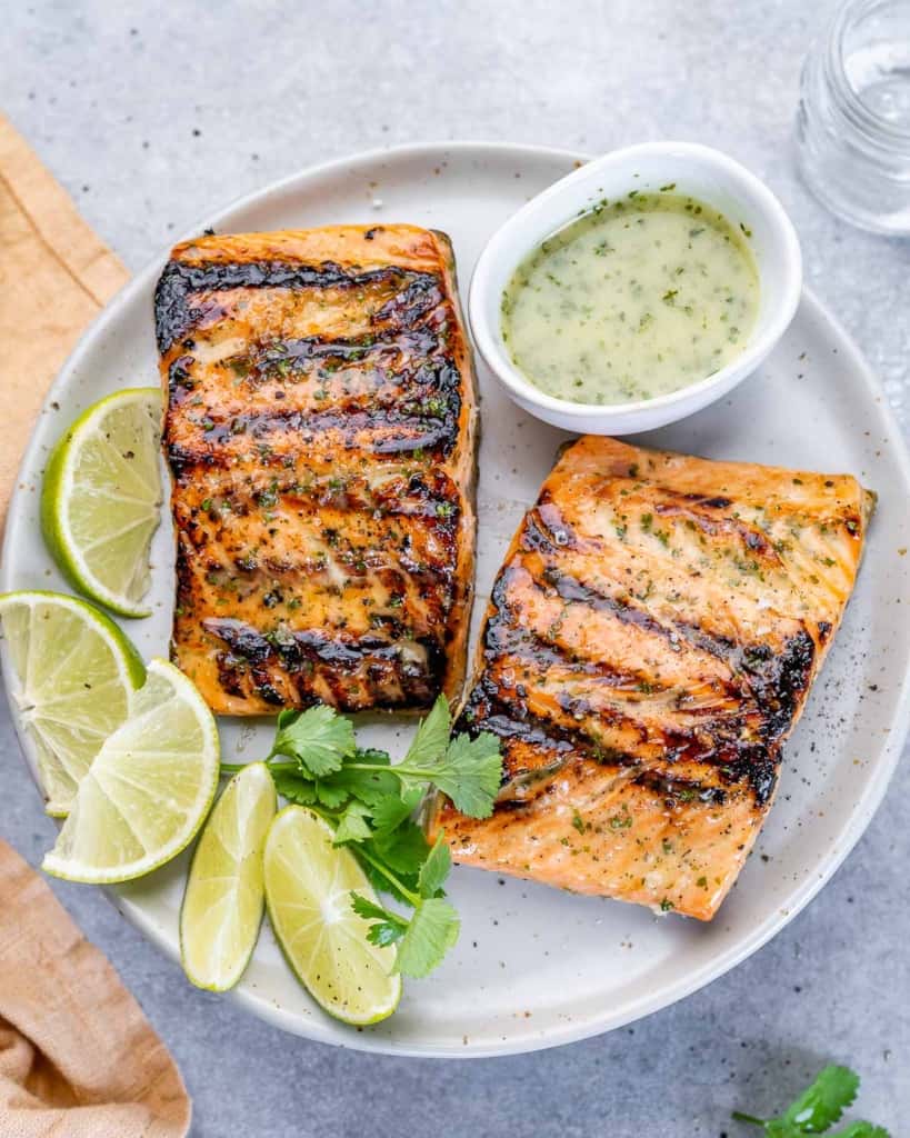 top view of two grilled salmon filets on a white plate with lime garnishes and cilantro lime drizzle in a small bowl