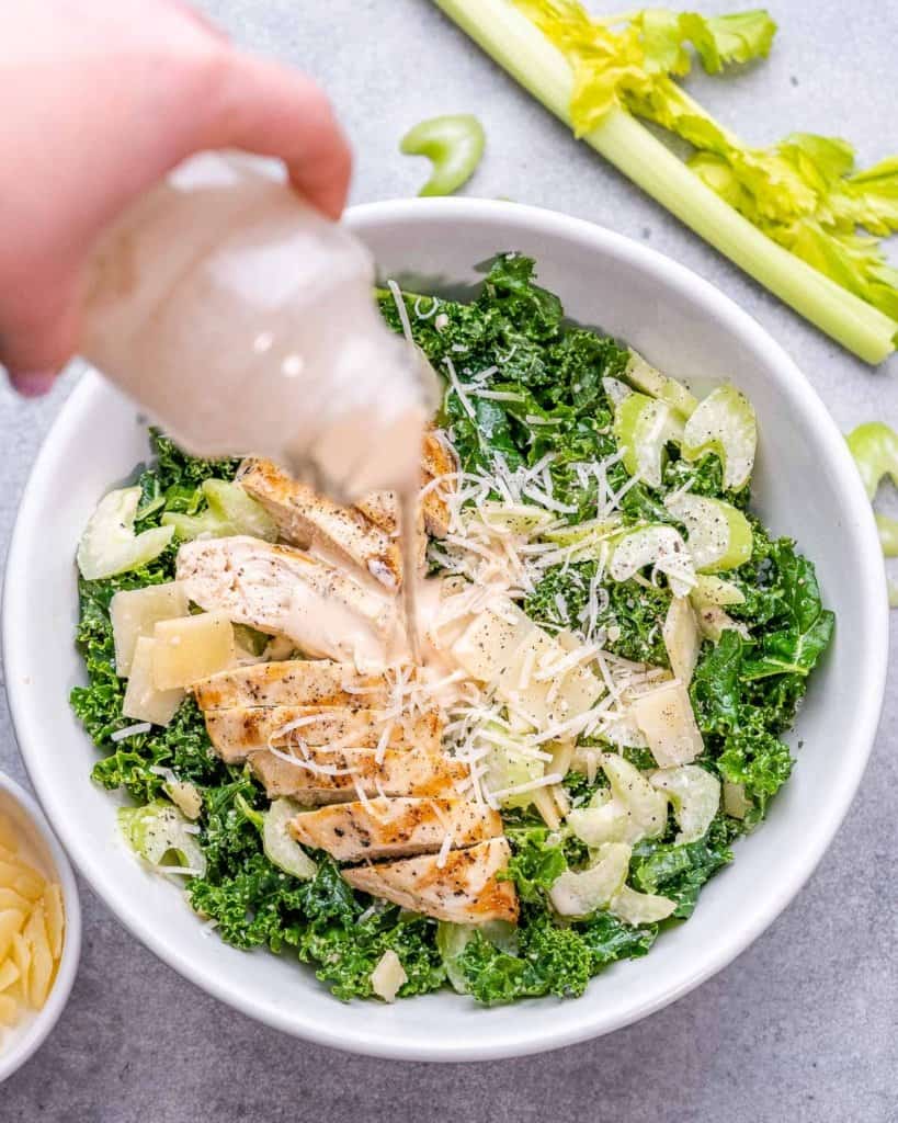 salad dressing in a bottle being poured over chicken kale salad in a white bowl