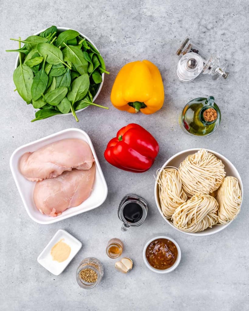 Ingredients on a grey table (chicken, spinach, egg noodles, peppers, garlic, various condiments, spices, and oils).