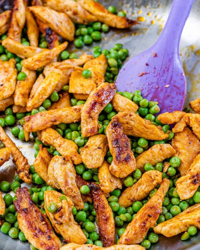Cooked chicken with peas in a stainless steel pan with a purple spatula.
