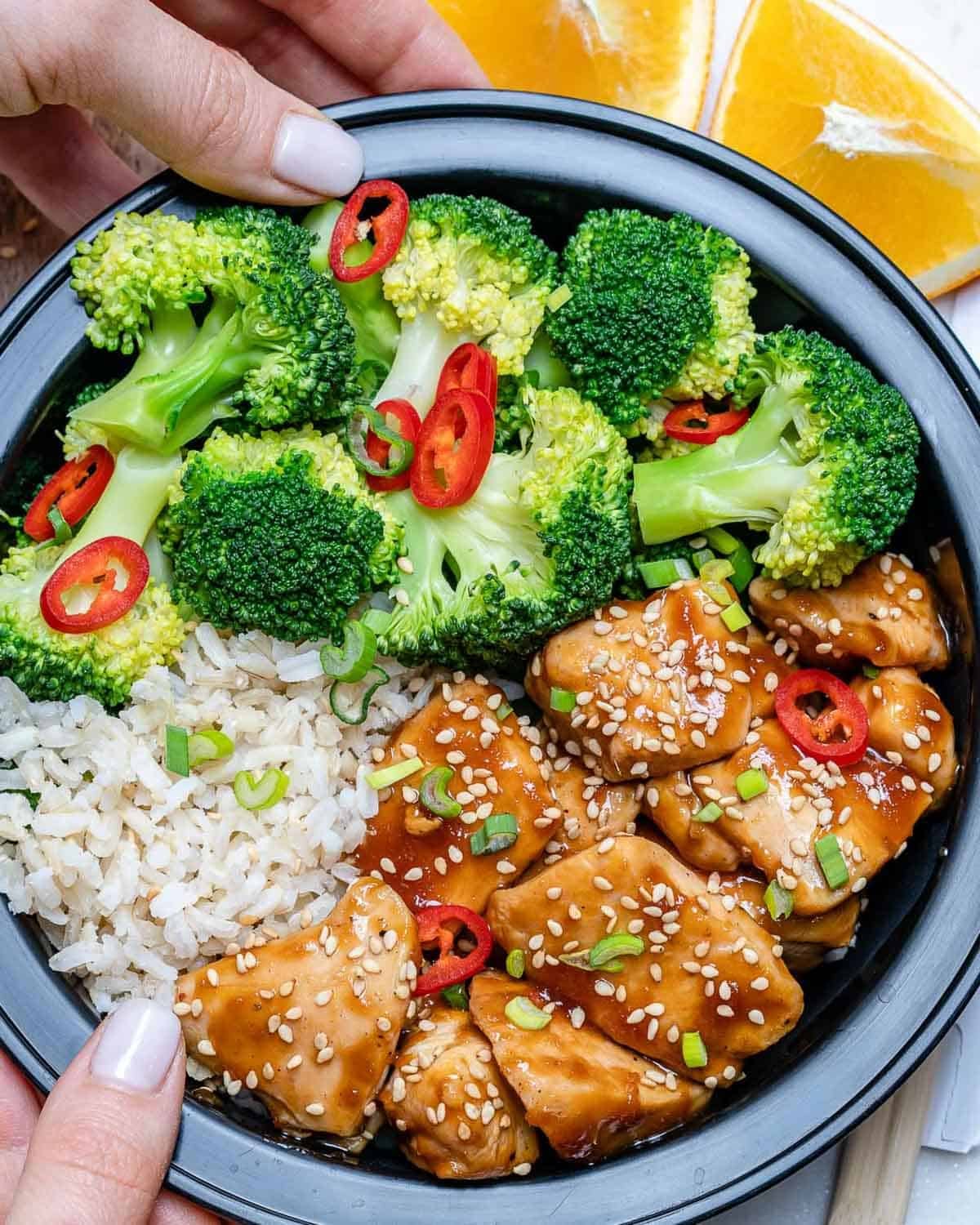 hand holding a bowl with chicken teriyaki, rice, and broccoli, in a meal prep bowl.