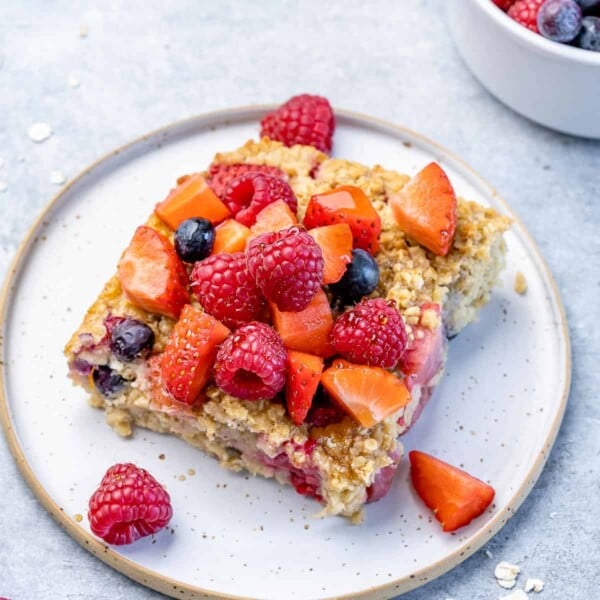 side shot of a slice of baked oatmeal on a white plate topped with berries and maple syrup
