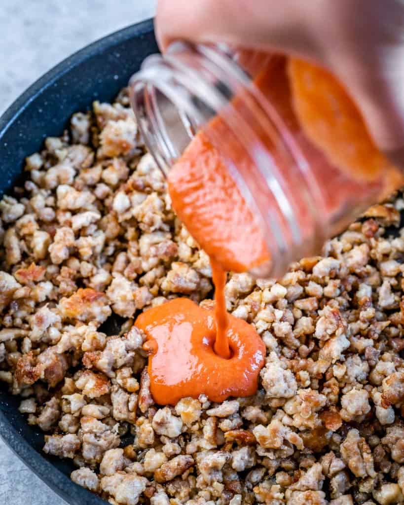 Adding buffalo sauce to ground chicken in a black pan.