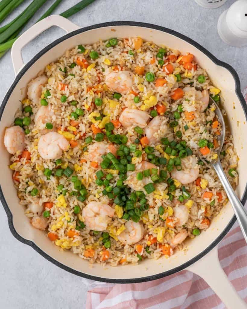Adding green onions to Shrimp Fried Rice in a white pan with a silver spoon.