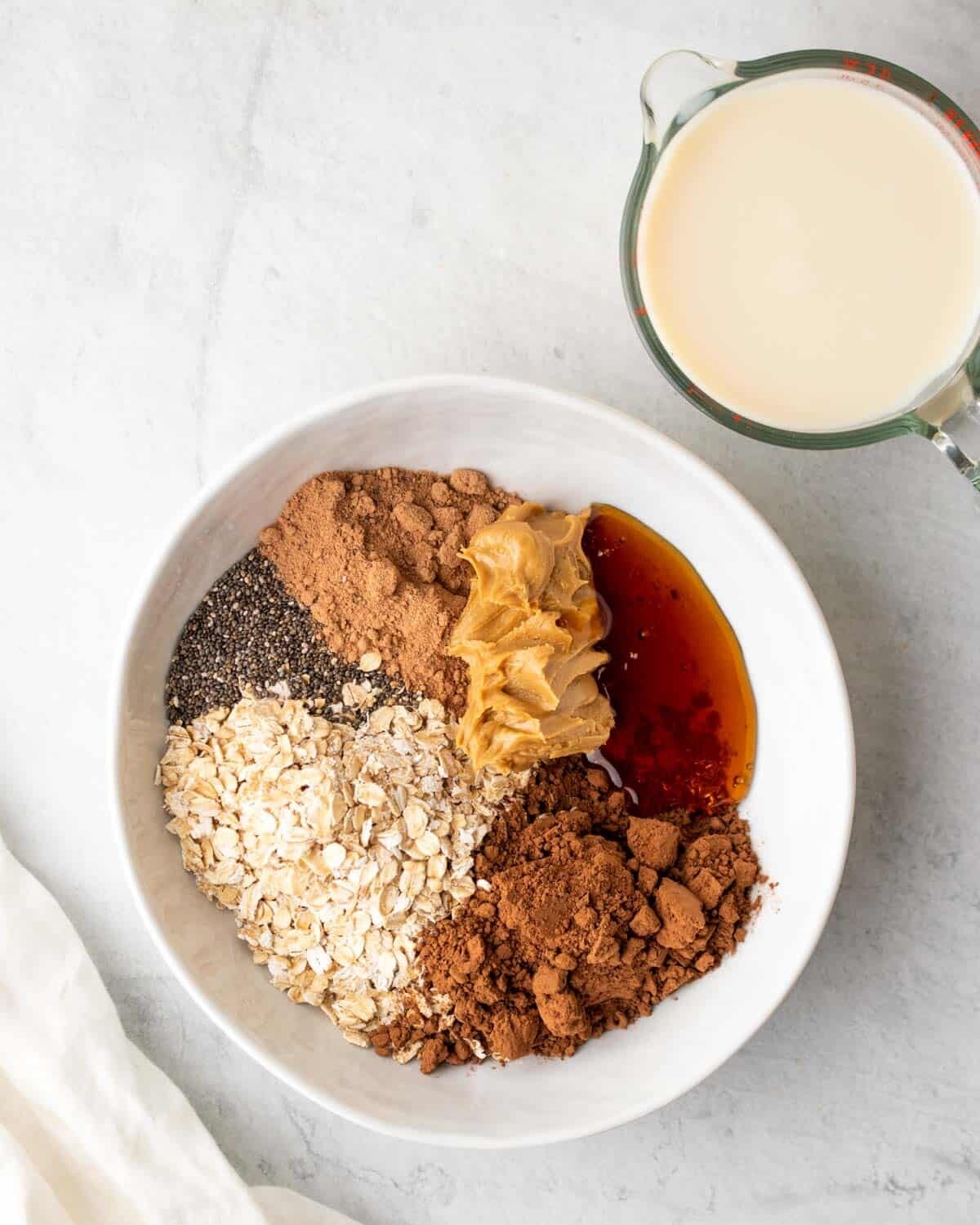 milk next to bowl with oats, chia seeds, protein powder, cocoa powder, maple syrup, and peanut butter.