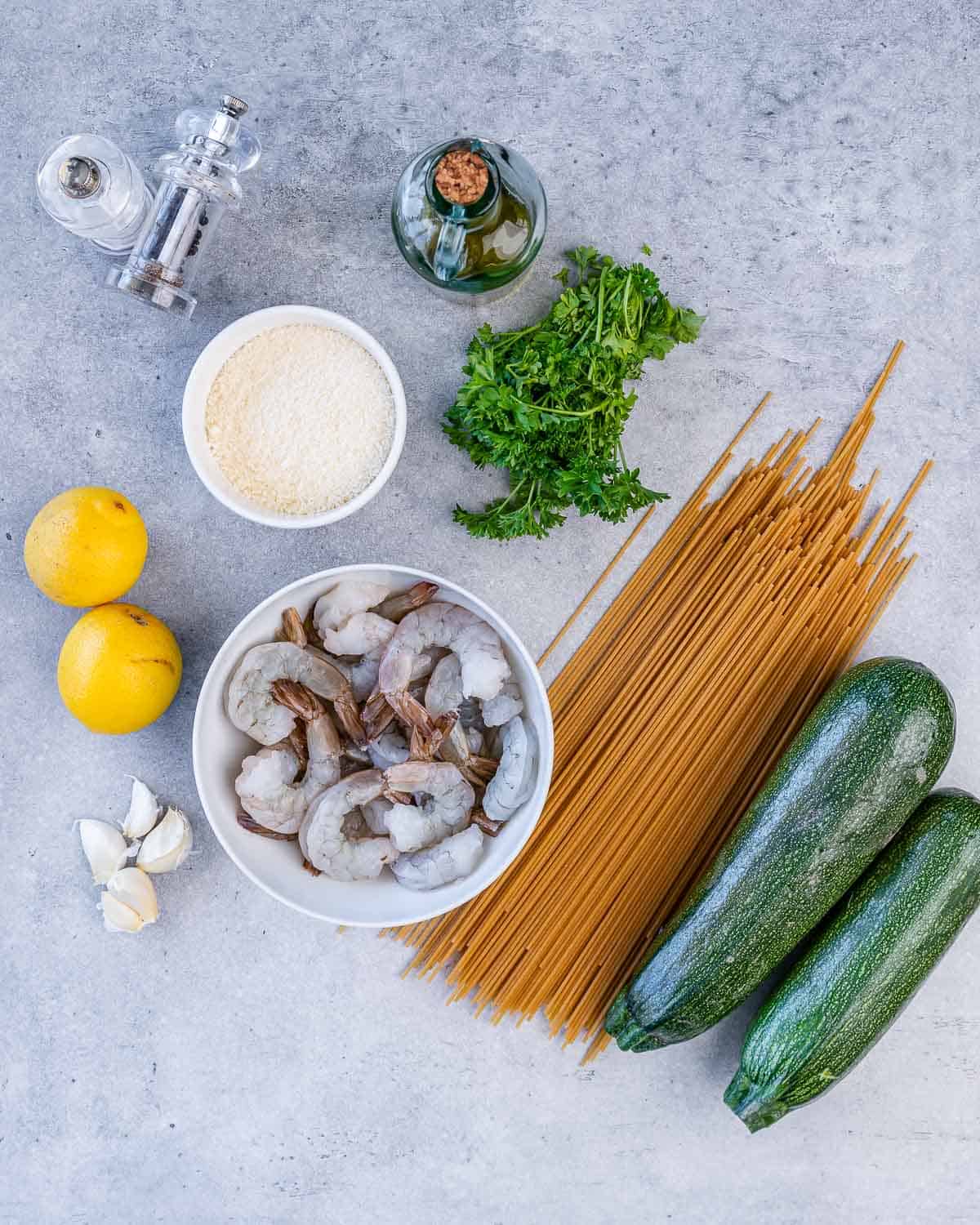 spaghetti, two zucchini, shrimp, parsley, olive oil, parmesan cheese, salt, pepper, two lemons, four garlic cloves on flat surface