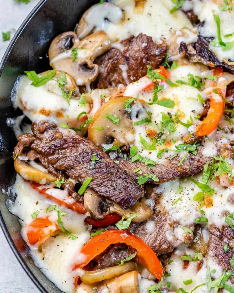 close up of cheesesteak in skillet with melted mozzarella and cilantro with bell peppers, onion, and mushrooms