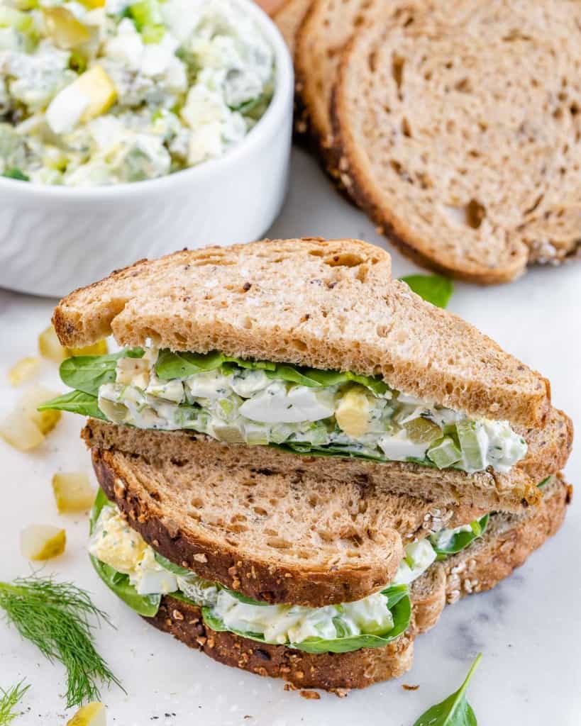 Side shot of an egg salad sandwich, halved and stacked on top of each other with ingredients in the background.