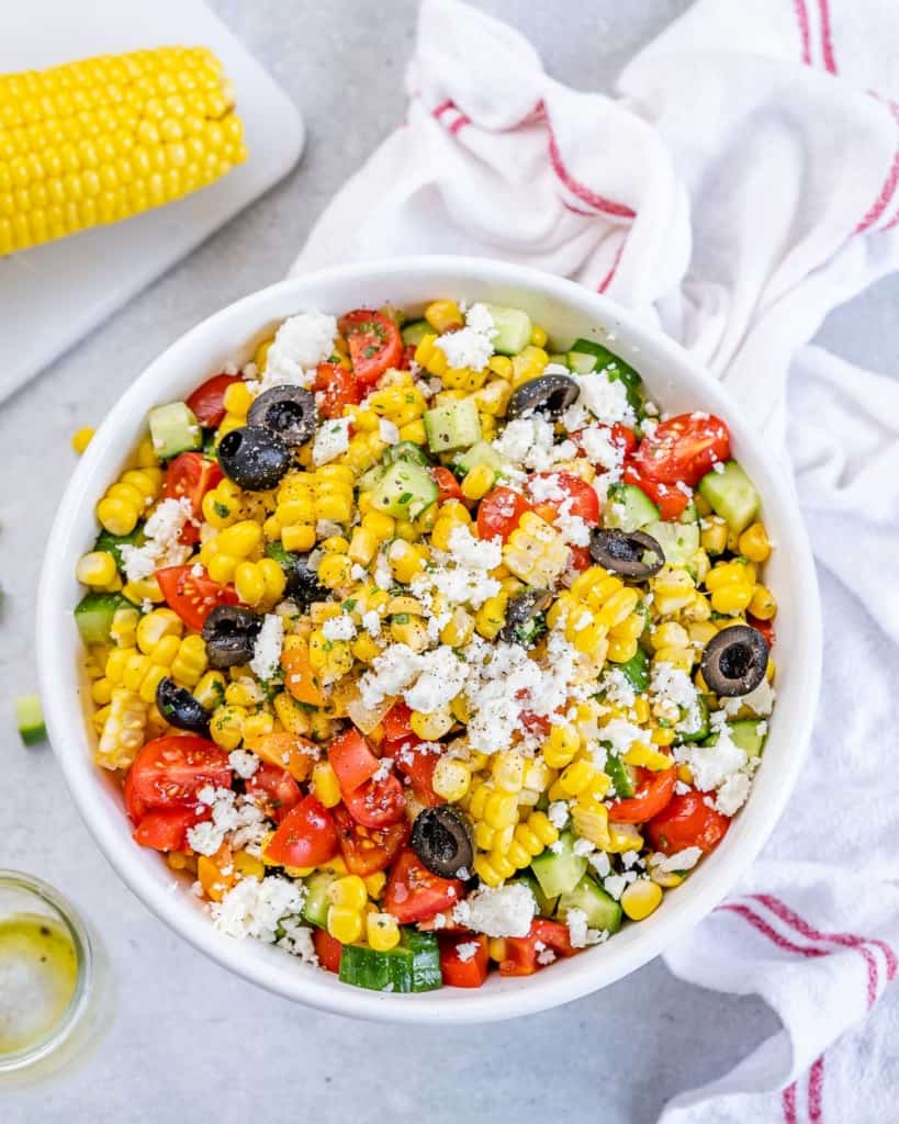 Overhead shot of summer corn salad in a white bowl surrounded by a cloth and ingredients on a table.
