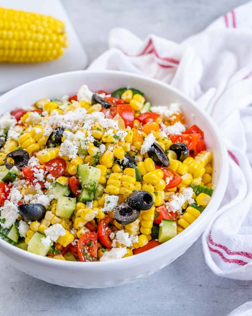 Side shot of summer corn salad in a white bowl surrounded by a cloth and ingredients on a table.