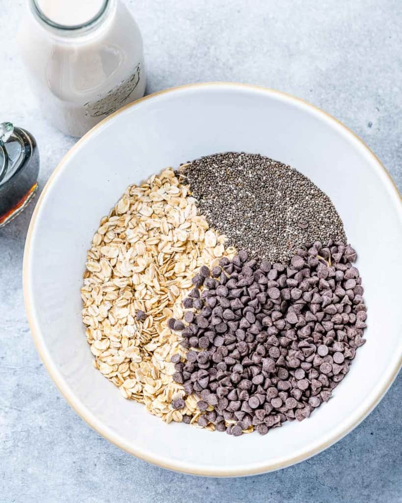 Oats, chocolate chips and chia seeds added to a white bowl.