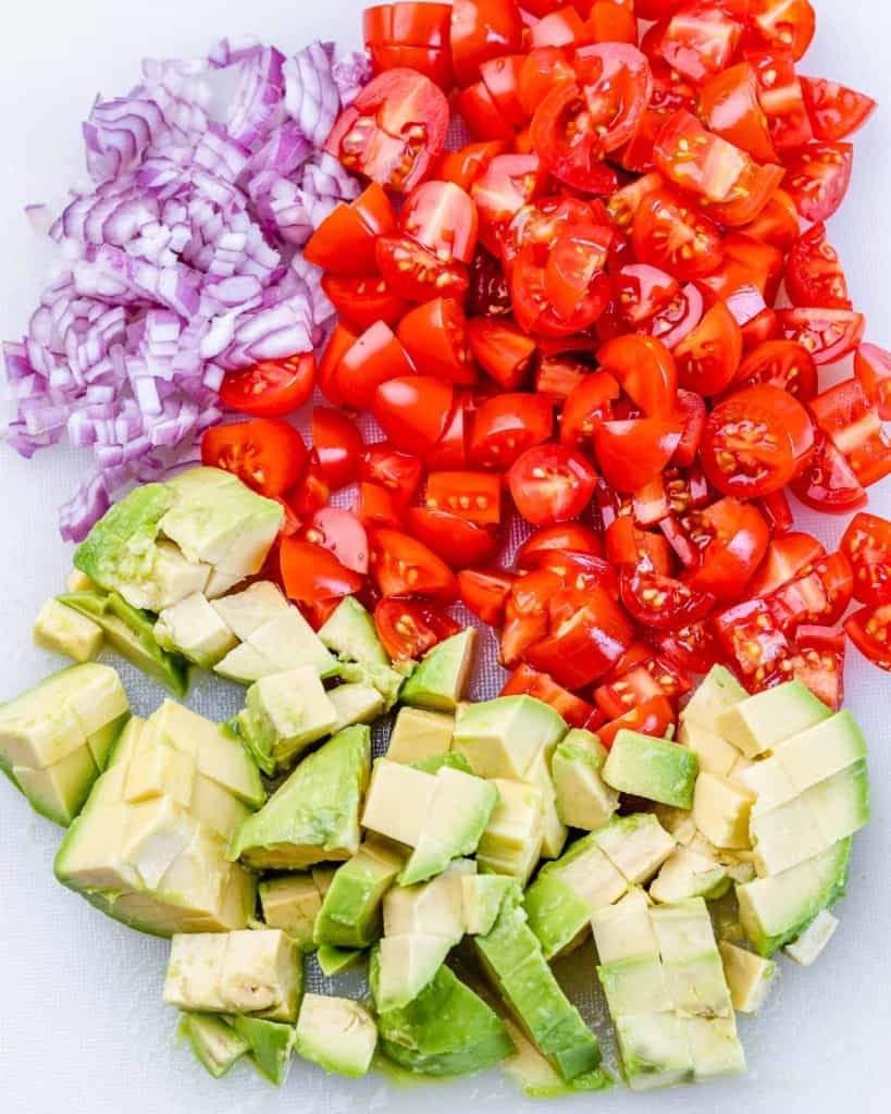 Chopped red onion, tomatoes and avocado on a cutting board.