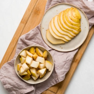 one plate of sliced pear next to a bowl of pear cubes with napkin and cutting board