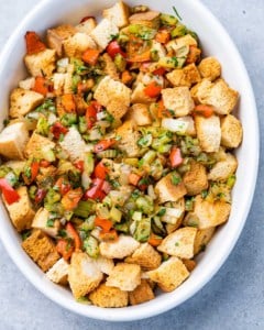 Tossing veggies with cubed bread in a white dish.