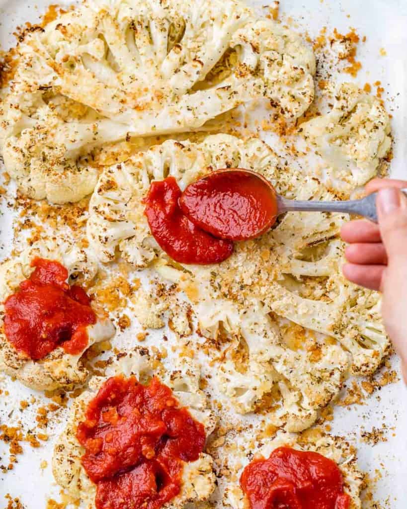 hand holding spoon and spreading marinara sauce over the baked cauliflower steaks