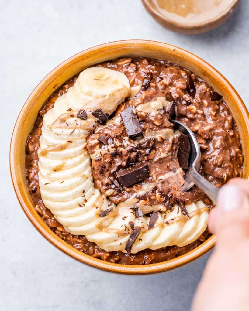 hand holding a spoon over chocolate oatmeal bowl topped with sliced bananas and shaved chocolate 