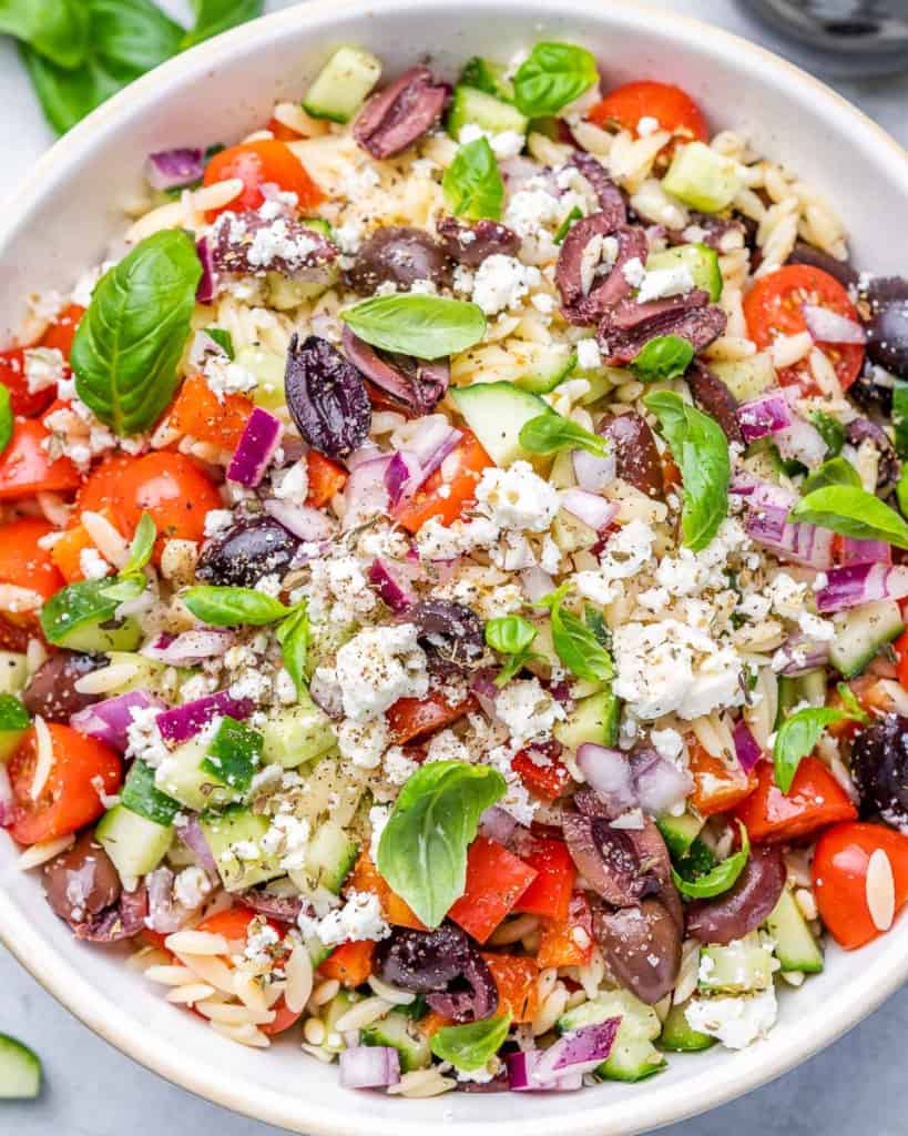 Orzo pasta salad with fresh veggies, basil and olives in a white bowl.