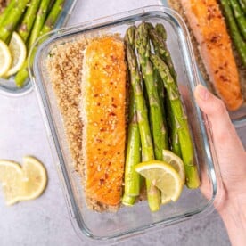 Woman's hand holding a glass container filled with salmon, asparagus, and quinoa.
