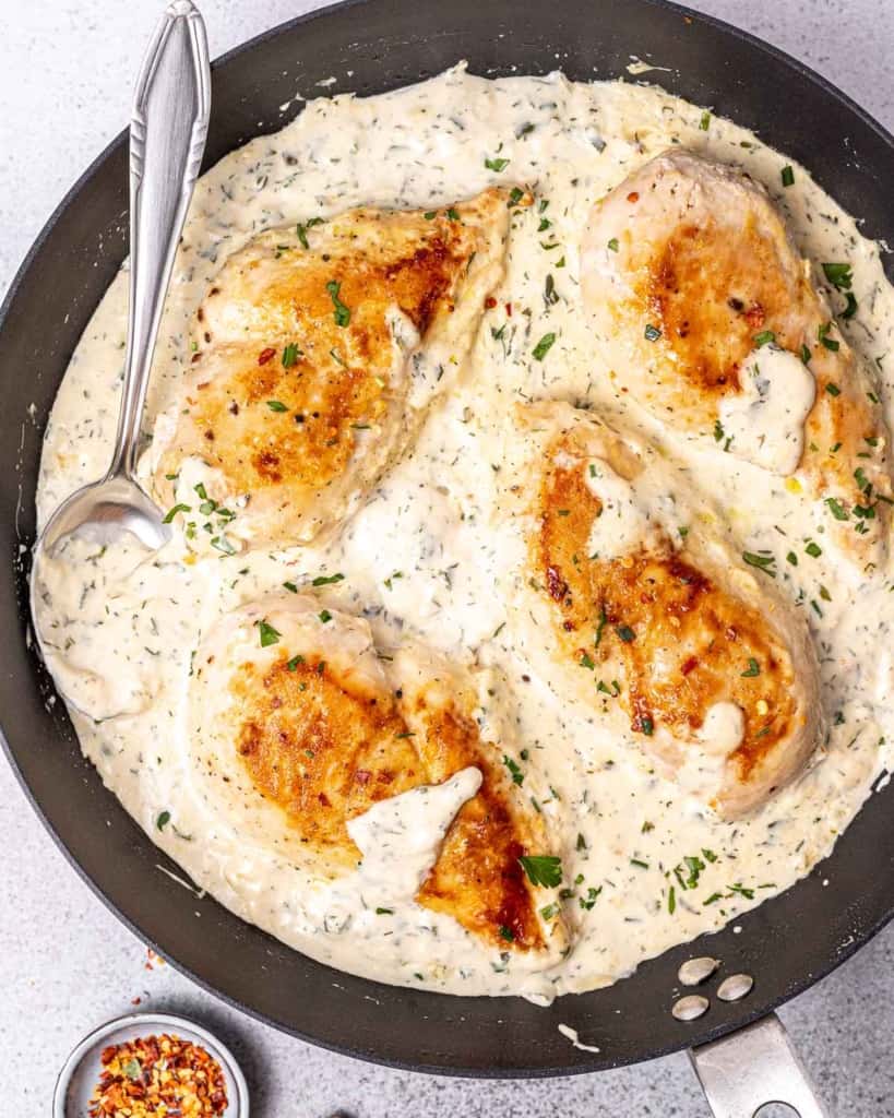 Spoon in a pan with chicken breasts cooked in a creamy white sauce.