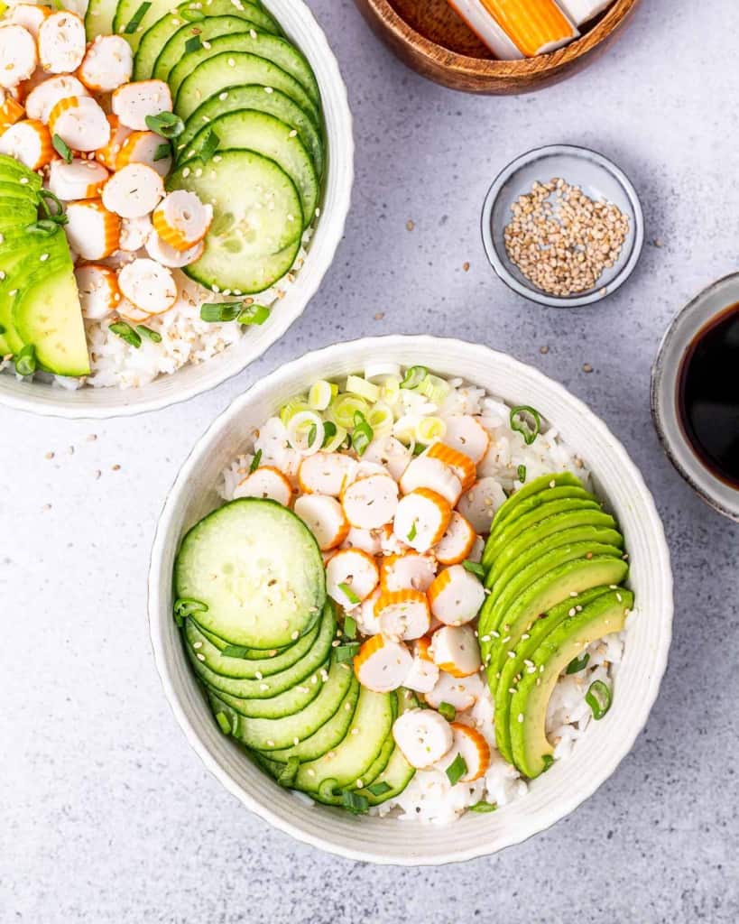 Assembling sushi bowls with rice, surimi, sliced cucumber and avocado.