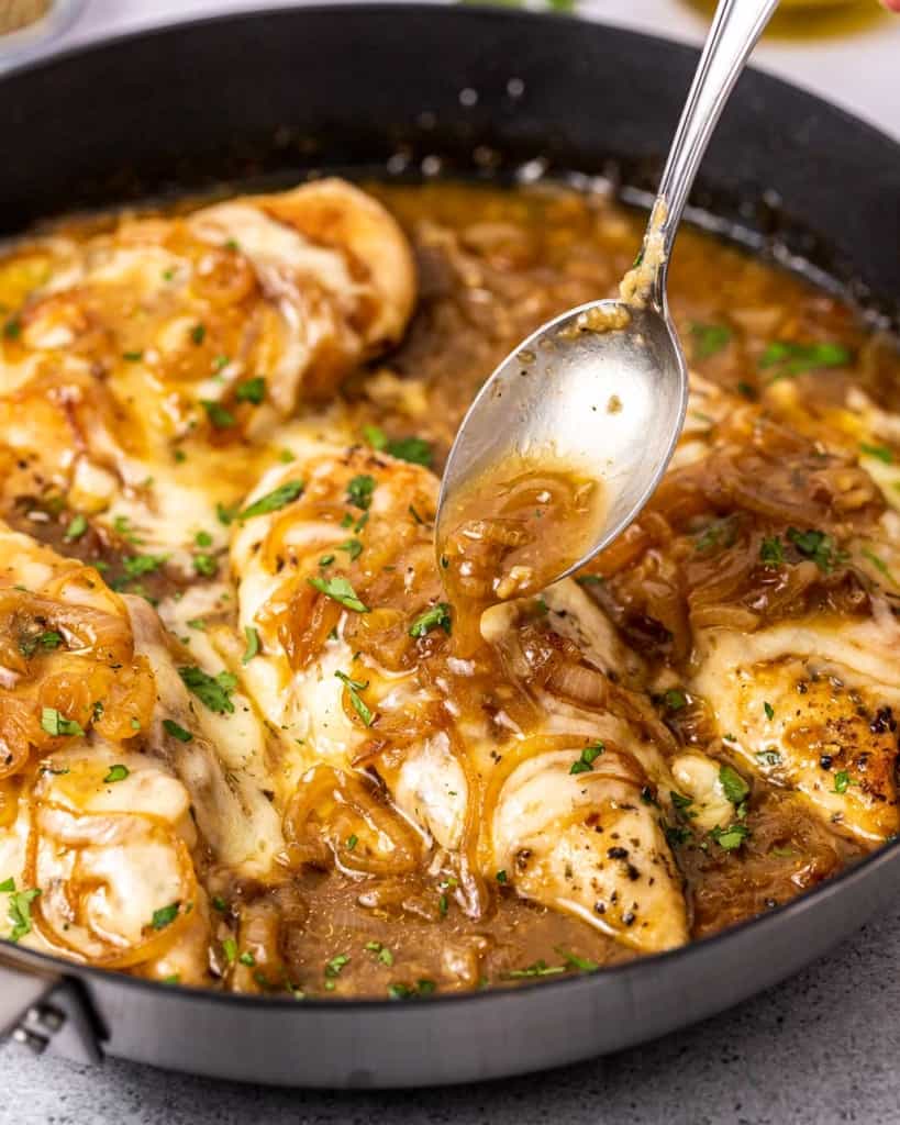 Spooning sauce over French onion chicken in a skillet.