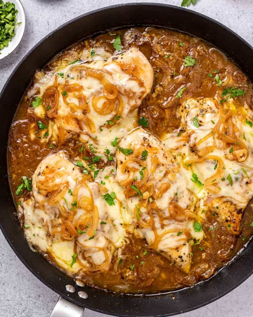 Top view of a black round skillet with 4 chicken breasts in an onion soup like sauce topped with melted cheese.