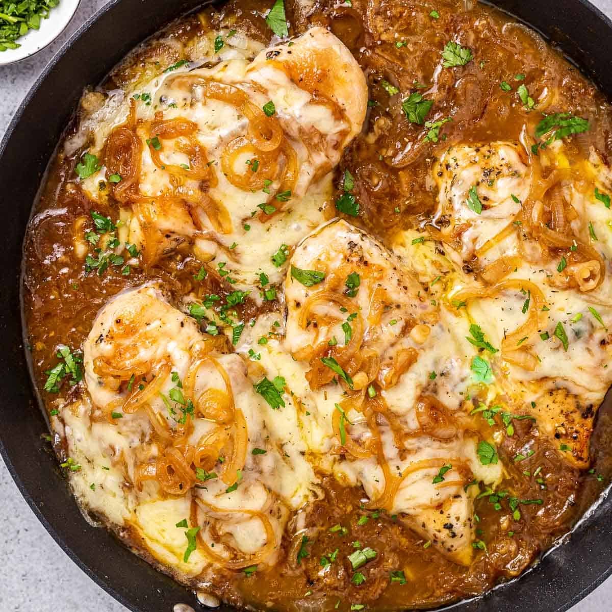 top view of a black round skillet with 4 chicken breasts in an onion soup like sauce topped with melted cheese