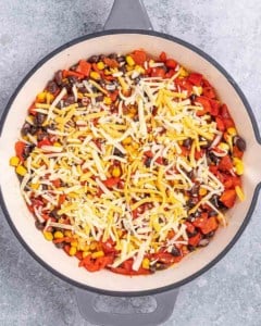 Overhead view of a skillet filled with shredded cheese, corn, black beans, and red peppers before baking.