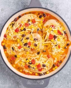 Skillet of chicken breasts covered in shredded cheese, ready to bake with a layer of beans, corn, and peppers underneath.