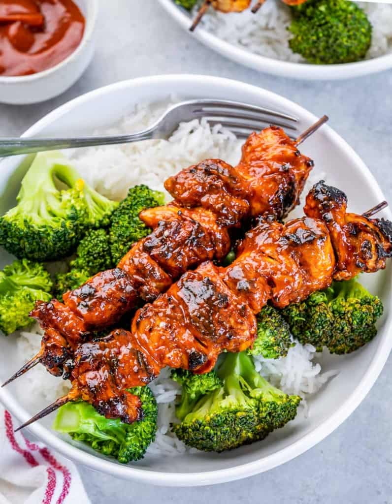 side shot of 2 chicken kabobs over rice and broccoli in a round white bowl