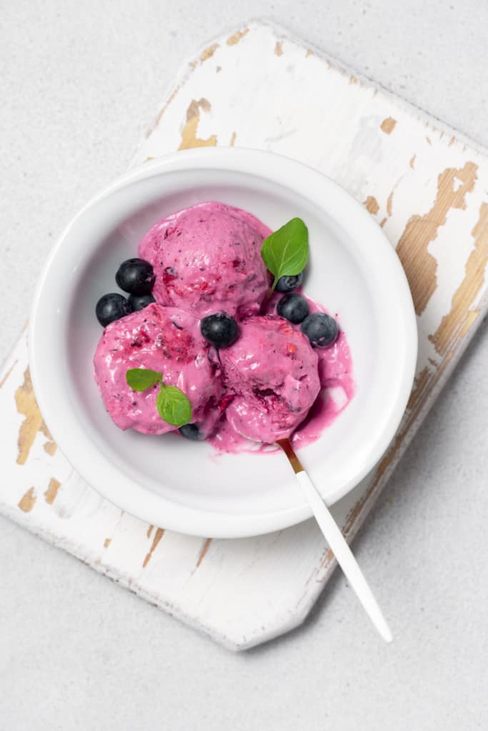 Scoops of purple colored ice cream in a white bowl with fresh berries and a spoon.