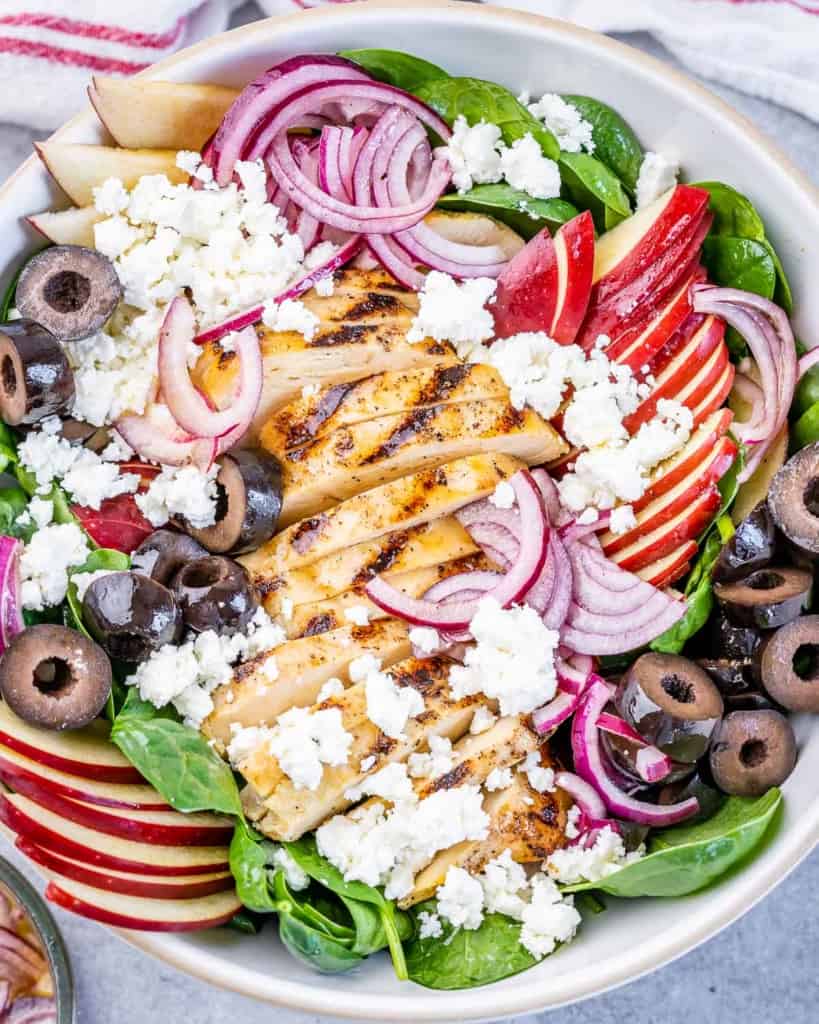 close up top view of spinach salad with grilled chicken, sliced apples, feta cheese, and olives 