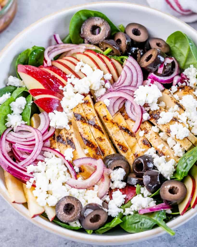 top view spinach salad with grilled chicken topped with sliced apples olives, and feta cheese