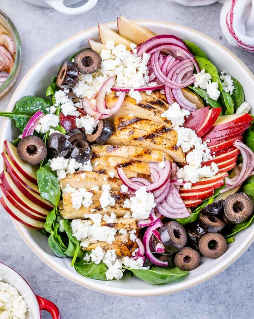 top  view salad in a white round bowl topped with grilled chicken, sliced onion, and feta cheese