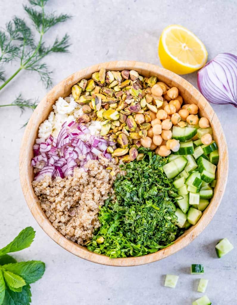Herbs, cucumber, quinoa, chickpeas and pistachios added to a large wooden bowl.